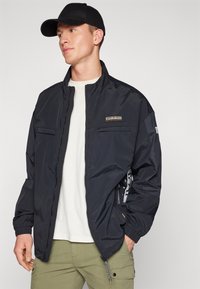 Chaqueta negra con cremallera y cuello alto, bolsillos laterales y puños elásticos. Llevada sobre una camiseta ligera y combinada con pantalones de cargo color oliva.