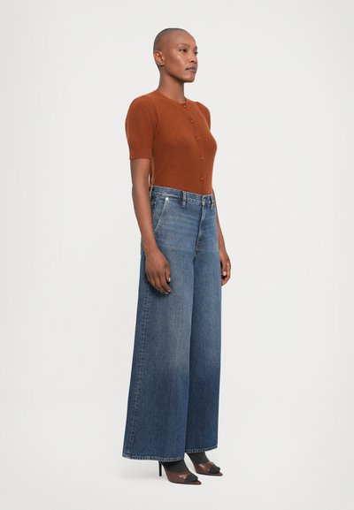 Maglia corta a maniche corte in maglia texturizzata marrone con bottoni, abbinata a jeans in denim blu a gamba larga. Scarpe nere con tacco completano l'outfit.