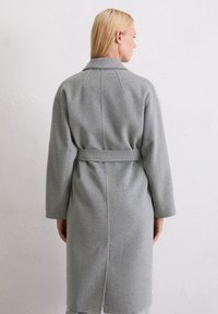 Grauer Wollmantel mit Gürtel, langen Ärmeln und Kragen. Besitzt einen geraden Schnitt, sichtbare Nahtdetails und einen glatten, strukturierten Stoff.