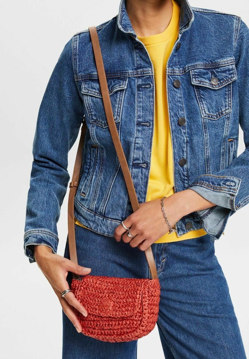 Borsa a tracolla intrecciata rossa con chiusura a pattina, caratterizzata da una finitura testurizzata e una tracolla in pelle marrone. Indossata con una giacca di jeans e un top giallo.