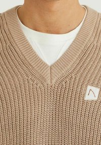 V-neck knitted sweater in light beige with a textured pattern. Features a small patch with a brown mountain outline label on the left side.