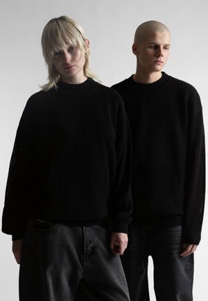 Dvaja mladí ľudia oblečení v čiernych textúrovaných svetroch a tmavých nohaviciach stoja vedľa seba na jednoduchom svetlom pozadí.
