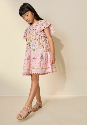 Young girl wearing a pink floral dress and light pink sandals, standing against a plain beige wall, looking down thoughtfully.