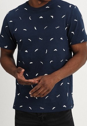 Mężczyzna ubrany w granatową koszulkę z rozproszonymi białymi wzorami wąsów, ręce swobodnie splecione na wysokości talii, na jednolitym tle.