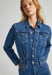 Džínový overal v středně modré barvě s dlouhými rukávy, zapínáním na knoflíky vpředu, dvěma hrudními kapsami a přiléhavým střihem. Obsahuje viditelné švy.