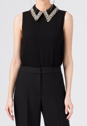 Haut noir sans manches avec col pointu orné de bijoux, associé à un pantalon noir taille haute ajusté porté par une personne debout.