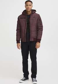 Bordeaux farvet jakke med hætte, lynlås foran og ribstrikkede manchetter. Sort sweater under jakken, parret med sorte bukser og sneakers.