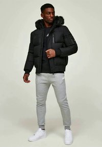 Homem a usar um casaco de puffer preto com capuz de pelagem, camisa preta, calças de moletom cinzas e ténis brancos, de pé contra um fundo simples.