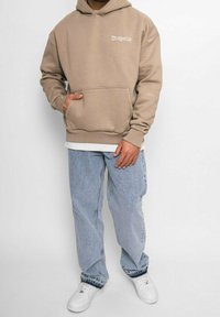 Sudadera beige con texto blanco "Dropsize," gran bolsillo tipo canguro, llevada sobre una camiseta blanca, combinada con jeans de mezclilla azul claro y zapatillas blancas.