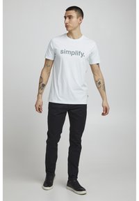 Hvid bomulds T-shirt med "simplify." trykt i gråt. Bæret med mørke bukser og sorte sneakers. Glat tekstur og afslappet pasform.