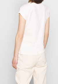 Camiseta de manga corta color beige claro, hecha de algodón, con cuello redondo y una silueta ligeramente ajustada. Diseño minimalista con líneas limpias.