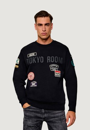 Ung man som bär en svart sweatshirt med texten "Tokyo Room" och flera färgglada lappar, kombinerat med mörka trasiga jeans, stående med ena handen i fickan.