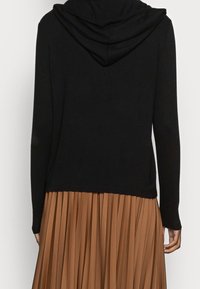 Černá mikina s kapucí vyrobená z měkkého materiálu. Má dlouhé rukávy a zkrácenou délku, doplněná o plisovanou hnědou sukni.