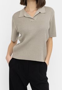 Beige geribbelde trui met korte mouwen, V-hals en een zachte textuur, gecombineerd met zwarte broeken.