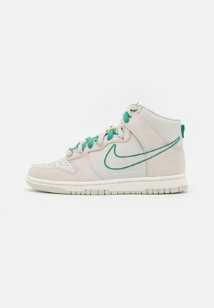 Beige højt snøresko fra Nike med grønt swoosh-omrids og snørebånd, synlige syninger og perforeringer på tåområdet på hvid baggrund.
