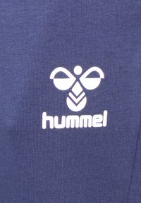 Dunkelblauer Stoff mit einem weißen Hummel-Logo. Das Logo enthält stilisierte Elemente, die an Flügel und ein Blättermuster erinnern.