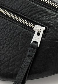 Zwarte leren crossbodytas met een gestructureerde afwerking, voorzien van twee zilveren ritsen en een platte leren band bevestigd aan een metalen bevestigingsstuk.