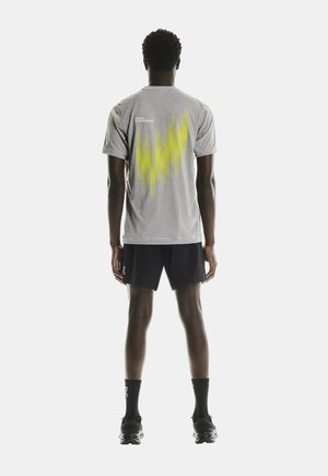 Graues Sport-T-Shirt mit gelbem abstraktem Muster auf dem Rücken, kombiniert mit schwarzen Shorts und Sportschuhen, von hinten betrachtet.