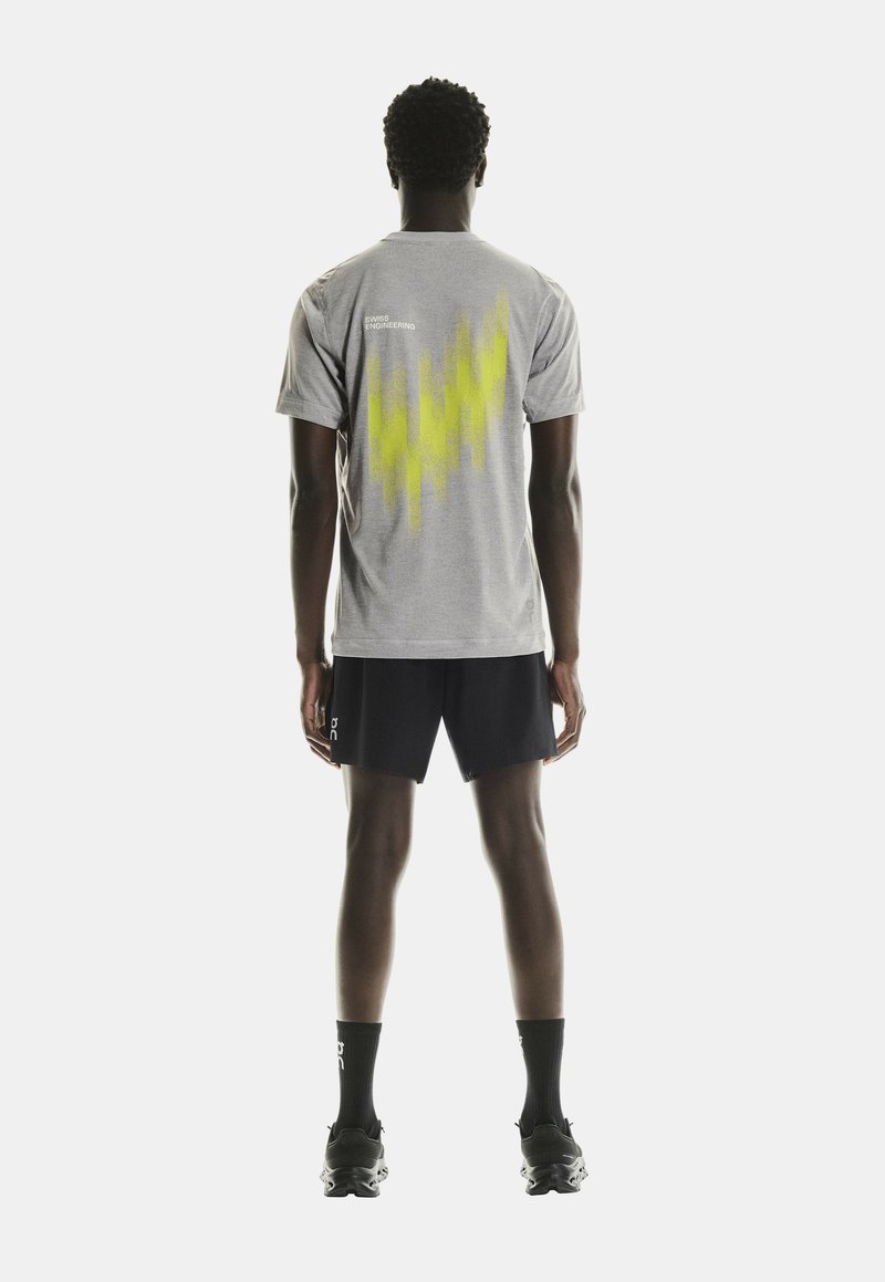 Grå atletisk t-shirt med ett gult abstrakt mönster på ryggen, matchad med svarta shorts och sportskor, sedd bakifrån.