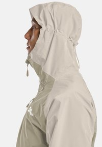 Profilo di una persona che indossa una giacca impermeabile bicolore beige con cappuccio e tasche con zip su uno sfondo semplice.