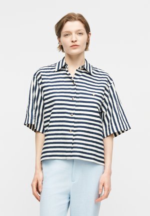 Άτομο με κοντά μαλλιά που φοράει πουκάμισο με οριζόντιες ρίγες σε ναυτικό μπλε και λευκό και ανοιχτό μπλε παντελόνι, μπροστά σε απλό φωτεινό φόντο.