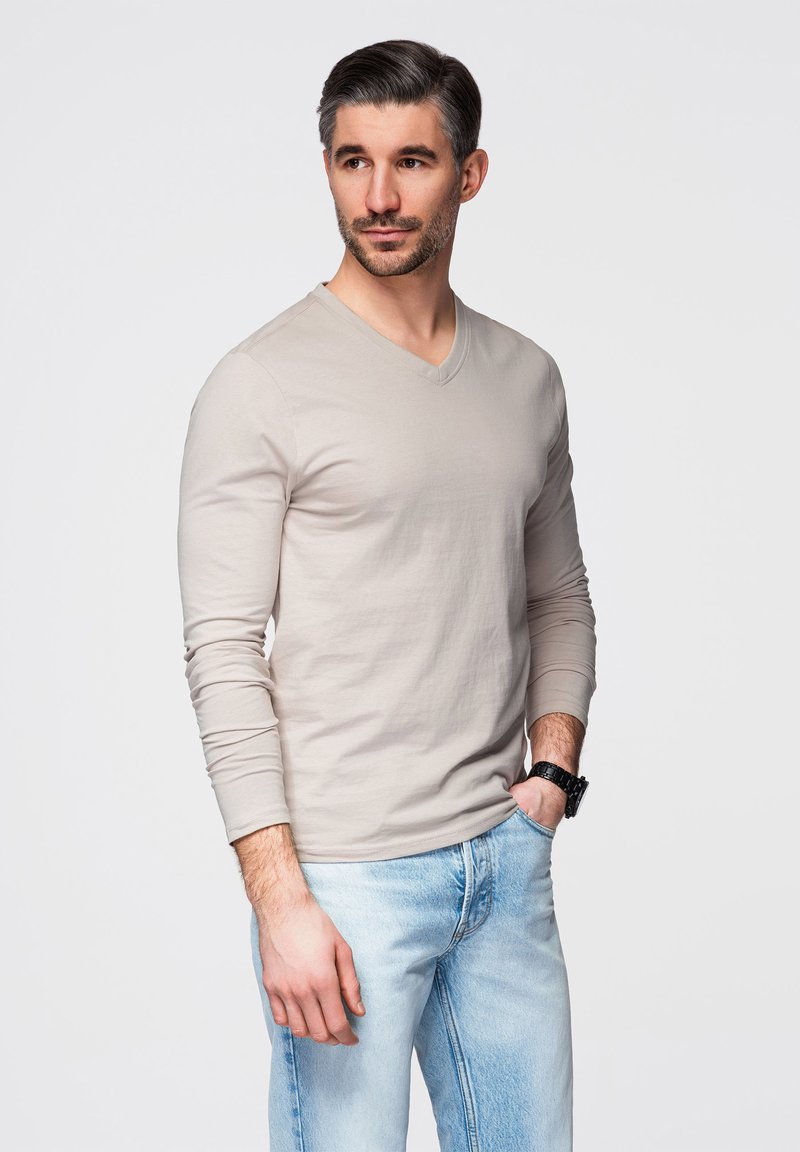 Hombre con cabello corto y oscuro y barba, que lleva una camisa beige de manga larga y vaqueros azul claro, de pie con una mano en el bolsillo.