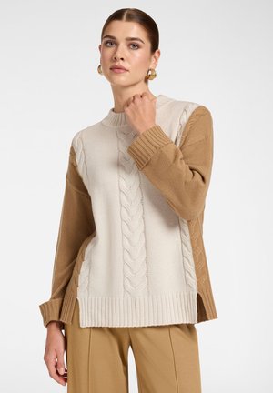 Vrouw met bruin haar die gouden oorringen, een beige en bruine kabelgebreide trui en beige broek draagt, poseert met hand bij haar hals.