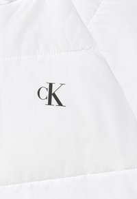 Bílá péřová bunda s prošívaným designem, hladkým povrchem a výrazným černým logem "cK" na levé straně.