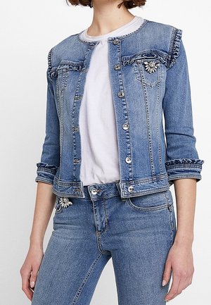 Kobieta ubrana w jasnoniebieską kurtkę jeansową z falbanami i kwiatowymi broszkami, w zestawie z dopasowanymi jeansami i białym t-shirtem.