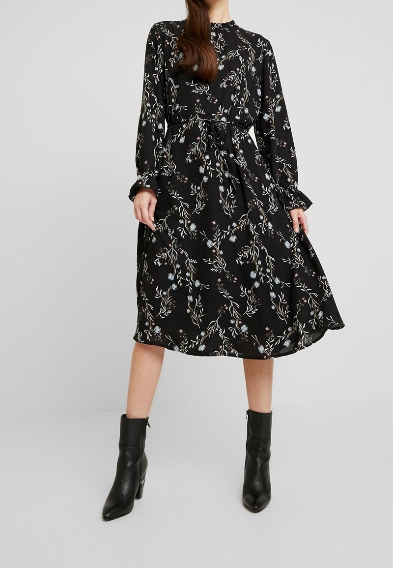 Femme portant une robe midi noire à imprimé floral, manches longues, poignets volantés, taille cintrée et bottines noires, debout sur un sol gris.