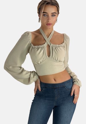 Lyse beige cropped top med lange, puffede ærmer, flæset halsudskæring og krydsede stropper; parret med mørkeblå tætsiddende jeans.