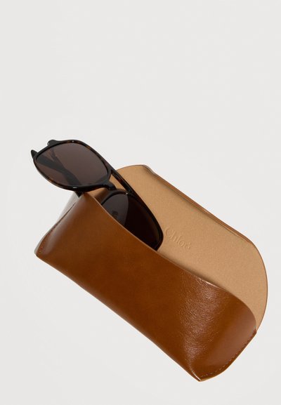 Σκούρα γυαλιά ηλίου με πλαίσιο από καβουρότσουκα, εν μέρει μέσα σε ανοιχτή καφέ δερμάτινη θήκη με μπεζ εσωτερικό.