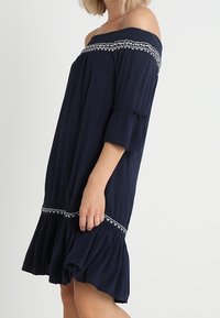 Robe bleu marine épaule dénudée en tissu léger, avec détails brodés et ourlet à volants. Manches courtes et coupe décontractée.