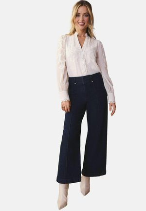 Blusa bianca a maniche lunghe con ricami floreali e dettagli con bottoni, abbinata a pantaloni larghi in denim scuro a vita alta e stivaletti beige.