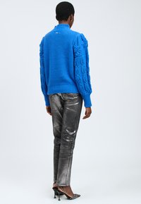 Maglione blu lavorato a maglia con motivo a trecce sulle maniche, col alto e bordo a coste; abbinato a pantaloni slim-fit argentati lucidi e décolleté a punta.