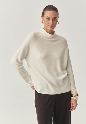 Heled kreemjas turtleneck-sviiter lõdva lõikega, ribakujulise tekstuuriga ja pikkade varrukatega, koos tumepruunide pükstega, millel on küljetaskud.