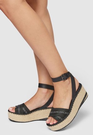 Sandalias de cuña negras con correas cruzadas, plataforma de yute texturizado, correa ajustable en el tobillo y marca visible en las correas.