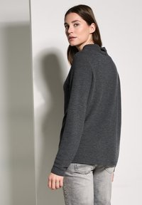 Grå randig långärmad topp med hög krage och texturerad design. Modellen bär ljusgrå jeans som visar en avslappnad passform.