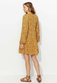Robe florale jaune moutarde à manches longues, taille cintrée et jupe évasée ; accompagnée de sandales brunes à lanières.