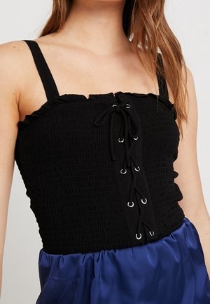 Femme portant un haut smocké noir sans manches avec un détail à lacets sur le devant et une jupe en satin bleue.