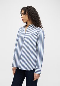 Γυναίκα με σγουρά μαλλιά που φοράει μπλε και λευκό πουκάμισο με κάθετες ρίγες και σκούρο παντελόνι, στέκεται πλαγιαστά μπροστά σε λευκό φόντο.