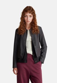 Giacca grigia abbottonata con un design aderente, due tasche frontali e tessuto testurizzato, abbinata a una maglietta bianca a coste e pantaloni bordeaux.