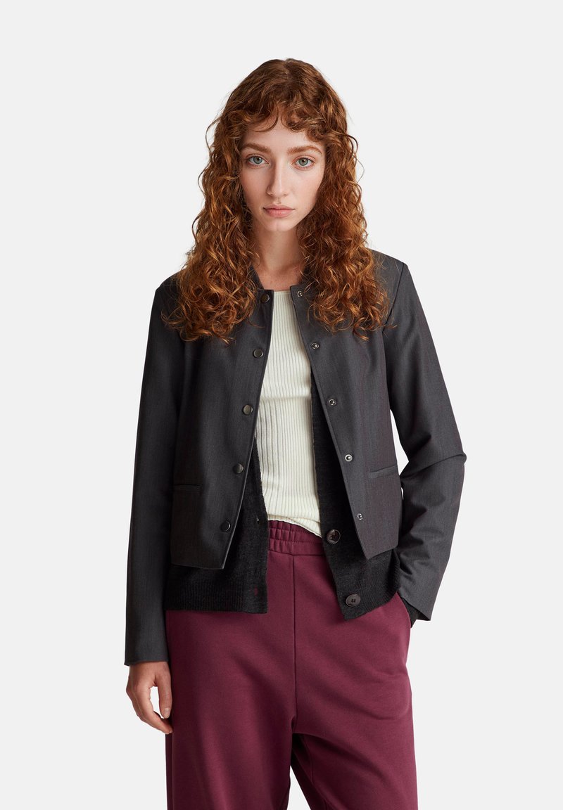 Giacca grigia abbottonata con un design aderente, due tasche frontali e tessuto testurizzato, abbinata a una maglietta bianca a coste e pantaloni bordeaux.
