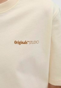 Kreminės spalvos medvilninė marškinė su apvalia apykakle. Priekyje yra mažas rudas "Originals™STUDIO" logotipas. Minkšta tekstūra.