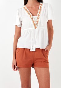 Blusa bianca con maniche corte e scollatura a V, con ricami floreali arancioni e nappine; abbinata a pantaloni corti color ruggine.