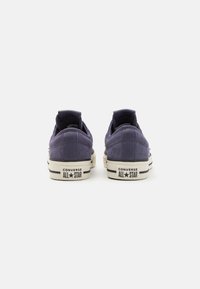 Sneakers low-top din suede violet, cu o talpă din cauciuc alb, având dungi negre și branding "Converse All Star" pe călcâi.