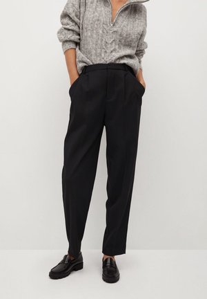 Personne portant un pantalon noir sur mesure, des mocassins noirs et un pull en maille torsadée gris avec une fermeture éclair mi-hauteur, les mains dans les poches.