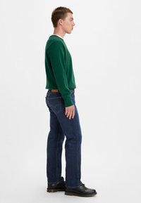 Grüner Pullover mit gerippten Bündchen, kombiniert mit blauen gerade geschnittenen Jeans. Schwarze Schuhe runden das Outfit ab. Einfarbiger weißer Hintergrund.