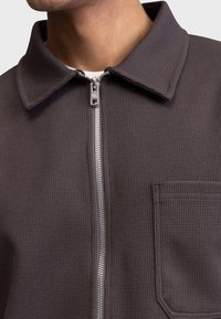 Chaqueta de color gris oscuro con cremallera, fabricada en un tejido texturizado y acanalado, con cuello puntiagudo y bolsillo en el pecho. Detalles en plateado en la cremallera y los herrajes.