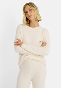 Pull beige clair en matière douce et duveteuse avec un col rond, des poignets côtelés et un petit détail en forme de cœur doré sur la poitrine. Associé à un bas assorti.